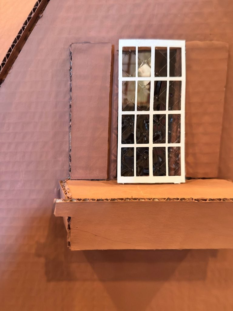 A window with white grid panes is set in a brown cardboard wall, flanked by two vertical panels over a small protruding ledge.