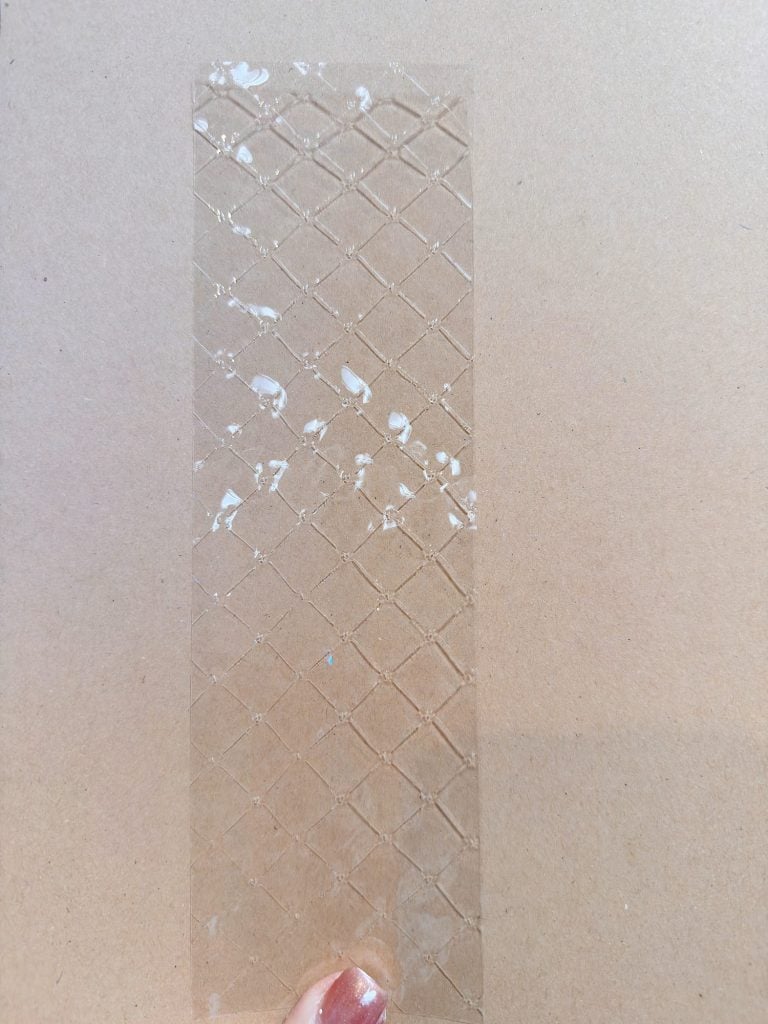 A hand holds a transparent, rectangular sheet with a crisscross pattern and some white spots, against a plain tan background.