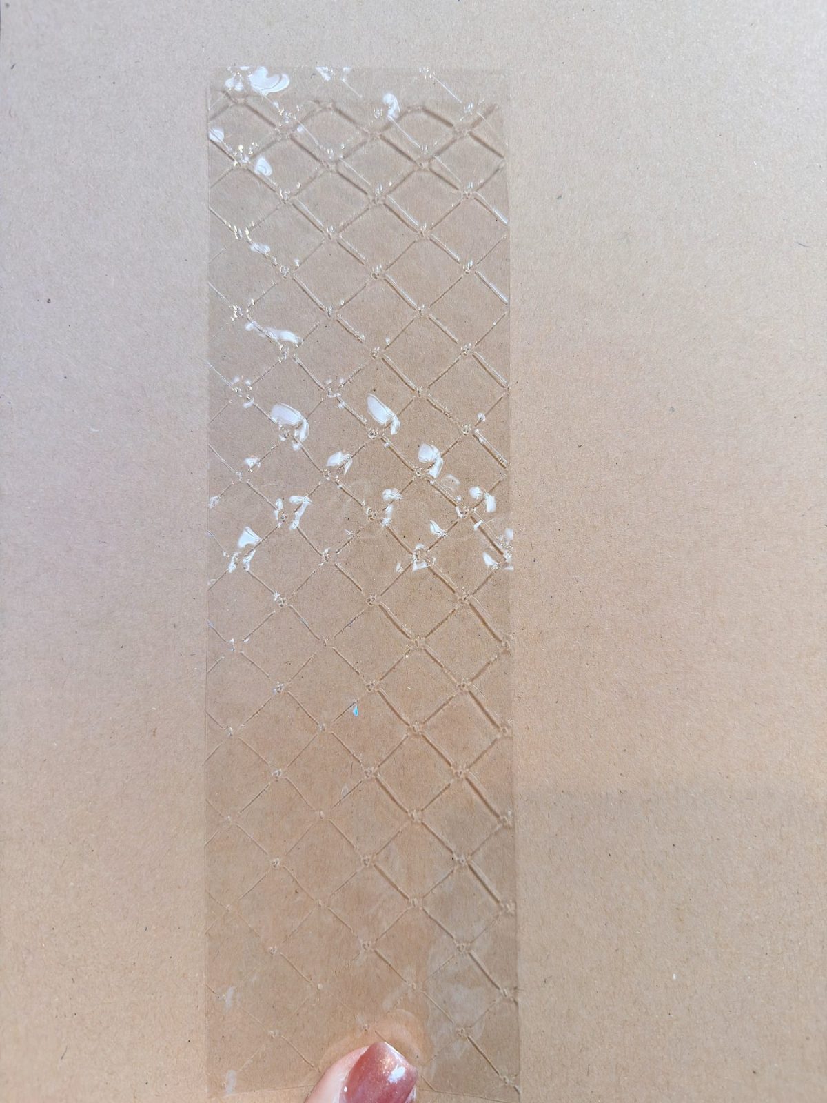 A hand holds a transparent, rectangular sheet with a crisscross pattern and some white spots, against a plain tan background.