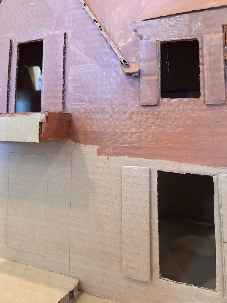 Close-up of a cardboard model of a building with rectangular window cutouts, some areas painted brown and others left unpainted.