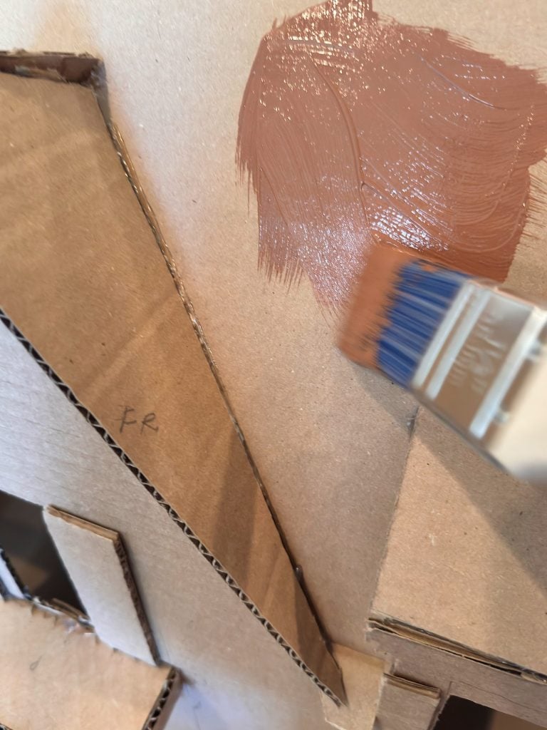 A paintbrush applies a coat of brown paint onto a piece of corrugated cardboard with other stacked cardboard shapes visible.