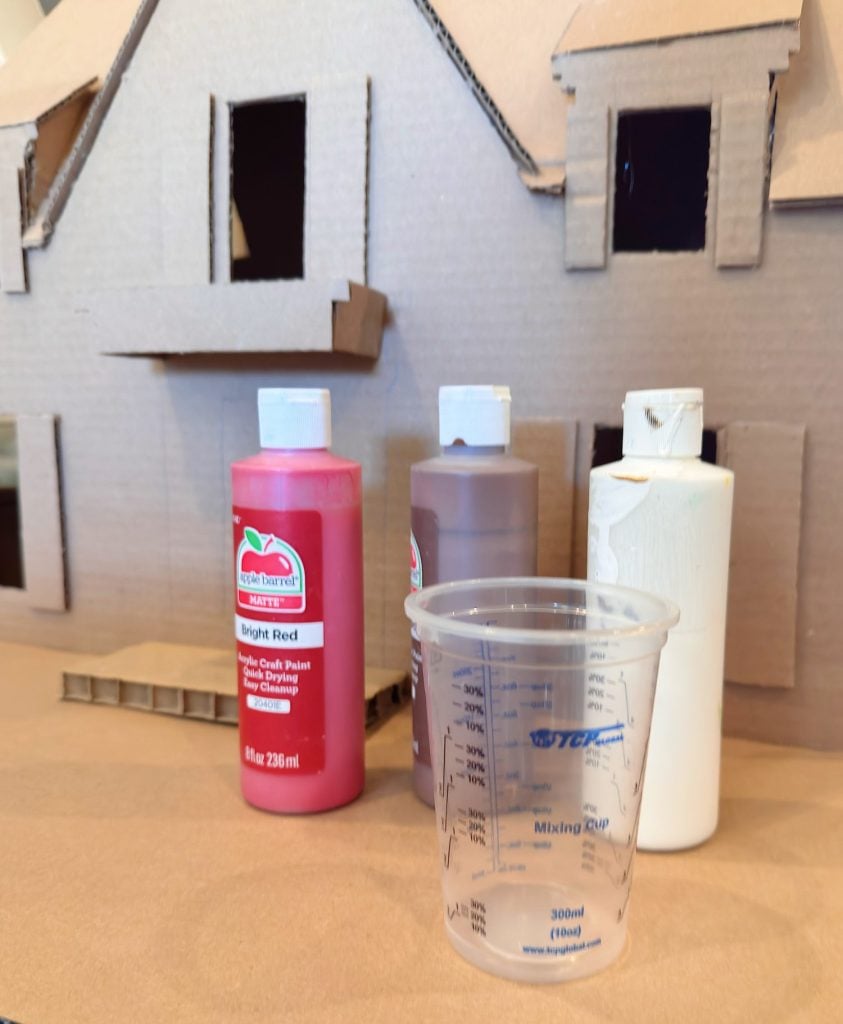 Three bottles of paint and a plastic mixing cup sit in front of an unfinished cardboard model house with cut-out windows.