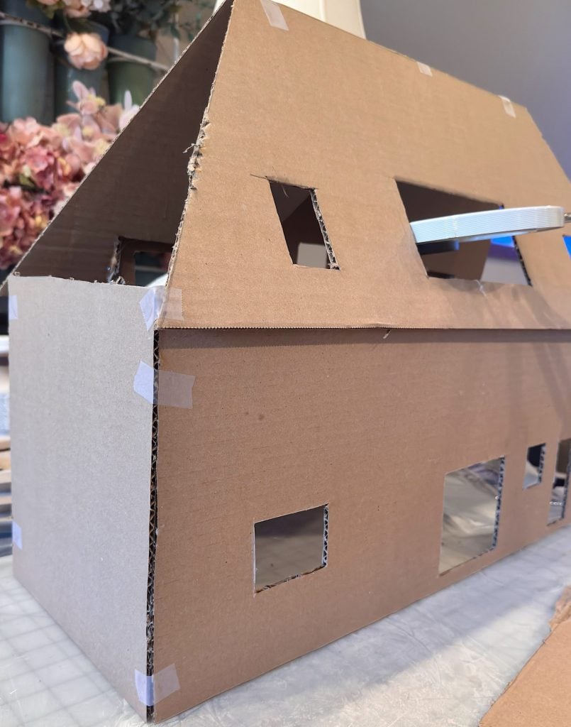 A cardboard model house with cut-out windows and tape securing the edges sits on a table, with flowers blurred in the background.