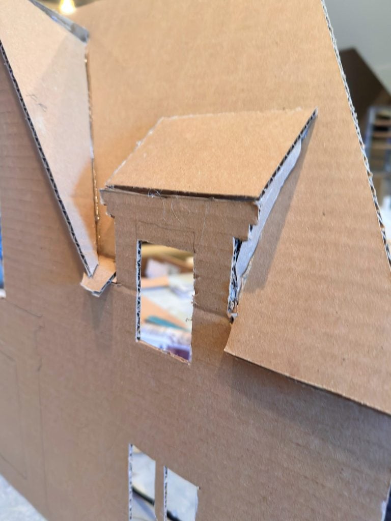 A close-up view of a cardboard model of a house, showing a dormer window cutout on the slanted roof.