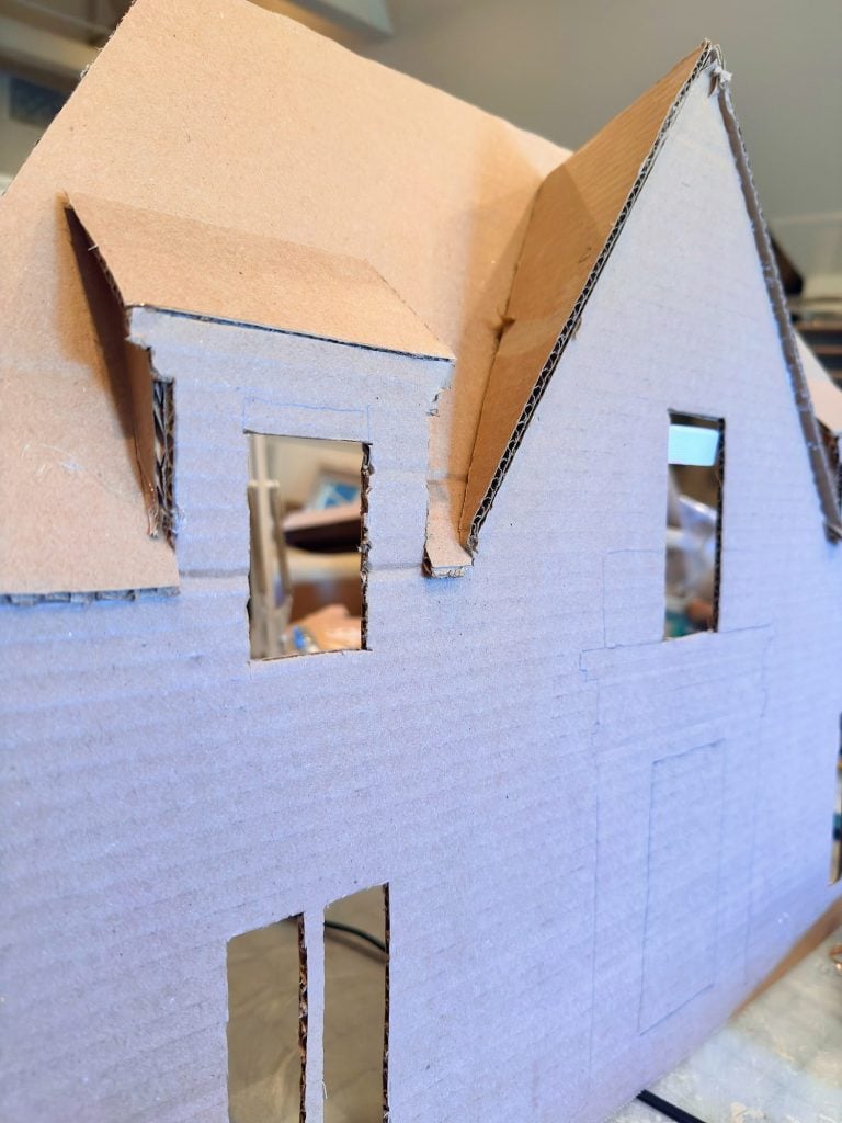 A close-up of a cardboard model house with cut-out windows and door, showing construction details and pencil lines marking features.