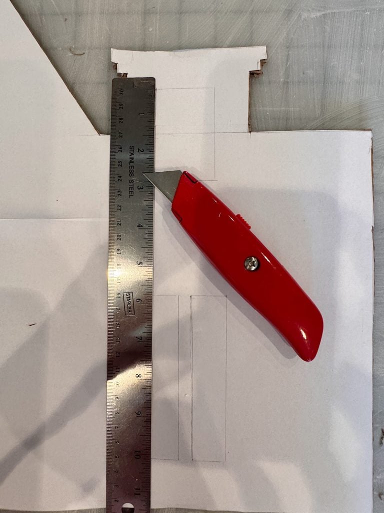A metal ruler and a red utility knife are placed on a piece of white cardboard marked with pencil lines and cut edges.