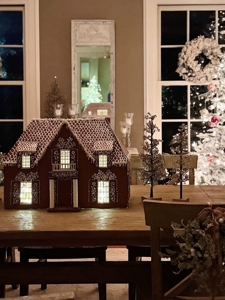 A gingerbread house with lit windows sits on a dining table, with small decorative trees nearby and a Christmas tree visible through the window in the background.