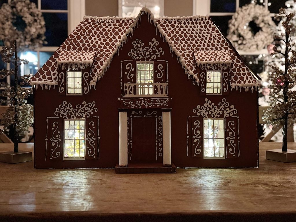 A gingerbread house decorated with white icing stands on a wooden surface, with its windows lit from within and festive decorations in the background.