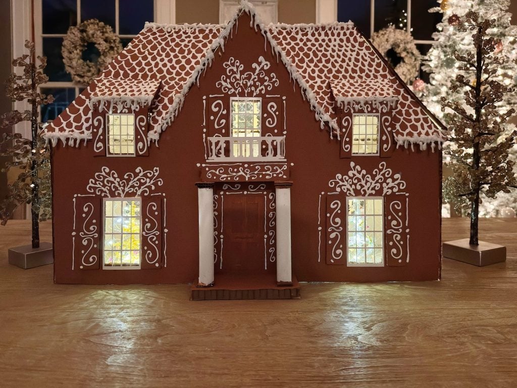 A gingerbread house with white icing decorations, lit windows, and a peaked roof is displayed on a wooden surface with holiday decor in the background.