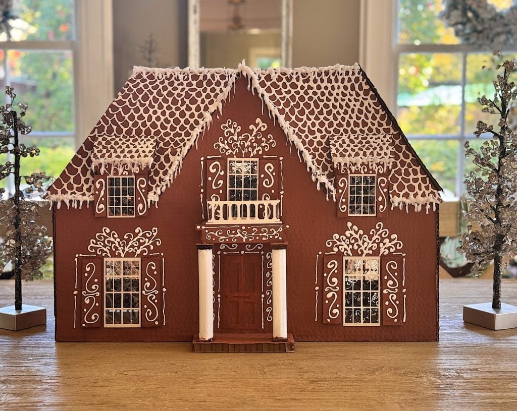 A large, detailed gingerbread house with white icing decorations sits on a wooden table, surrounded by small, silver holiday trees.