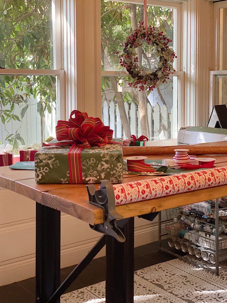 A table with wrapped holiday gifts, ribbon, and wrapping paper stands by windows decorated with a festive wreath.