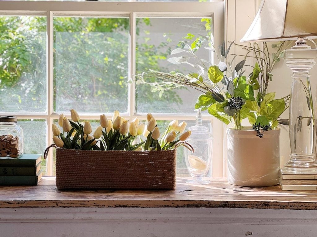 A windowsill with a wicker basket of white tulips, a ceramic vase with green and white foliage, a glass jar, books, and a table lamp; sunlight shines through the window&mdash;a perfect spot for reading or writing blog posts.