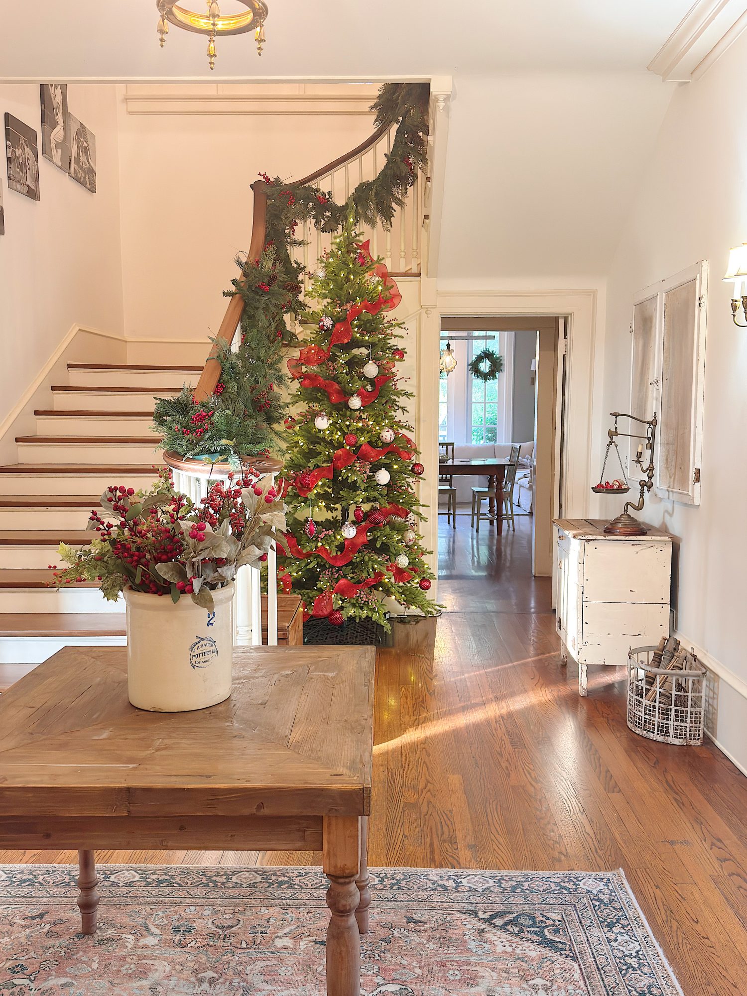 A staircase decorated with garland and a Christmas tree adorned with red and white ornaments stands in a bright hallway with wooden floors and holiday floral arrangements.