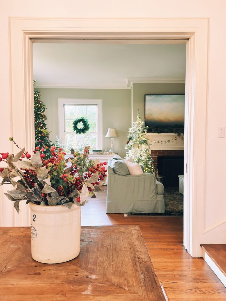 A living room decorated for the holidays with a wreath on the window, Christmas trees, and a vase with red berries on a wooden table in the foreground.