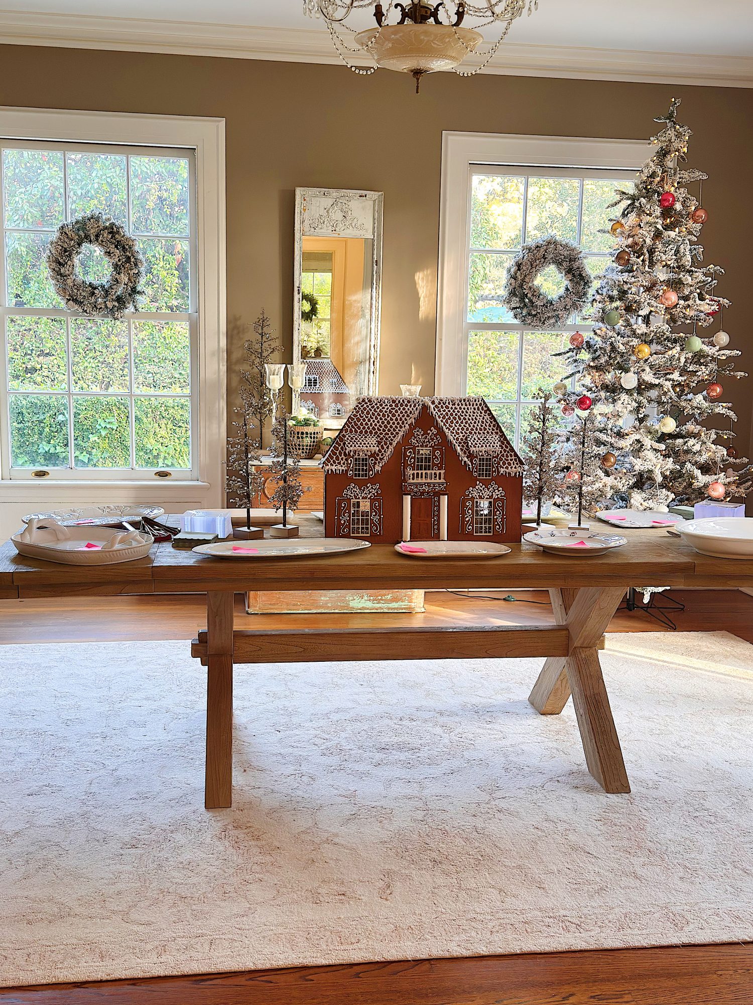 A decorated dining room with a gingerbread house centerpiece, a Christmas tree, wreaths on the windows, and festive table settings.
