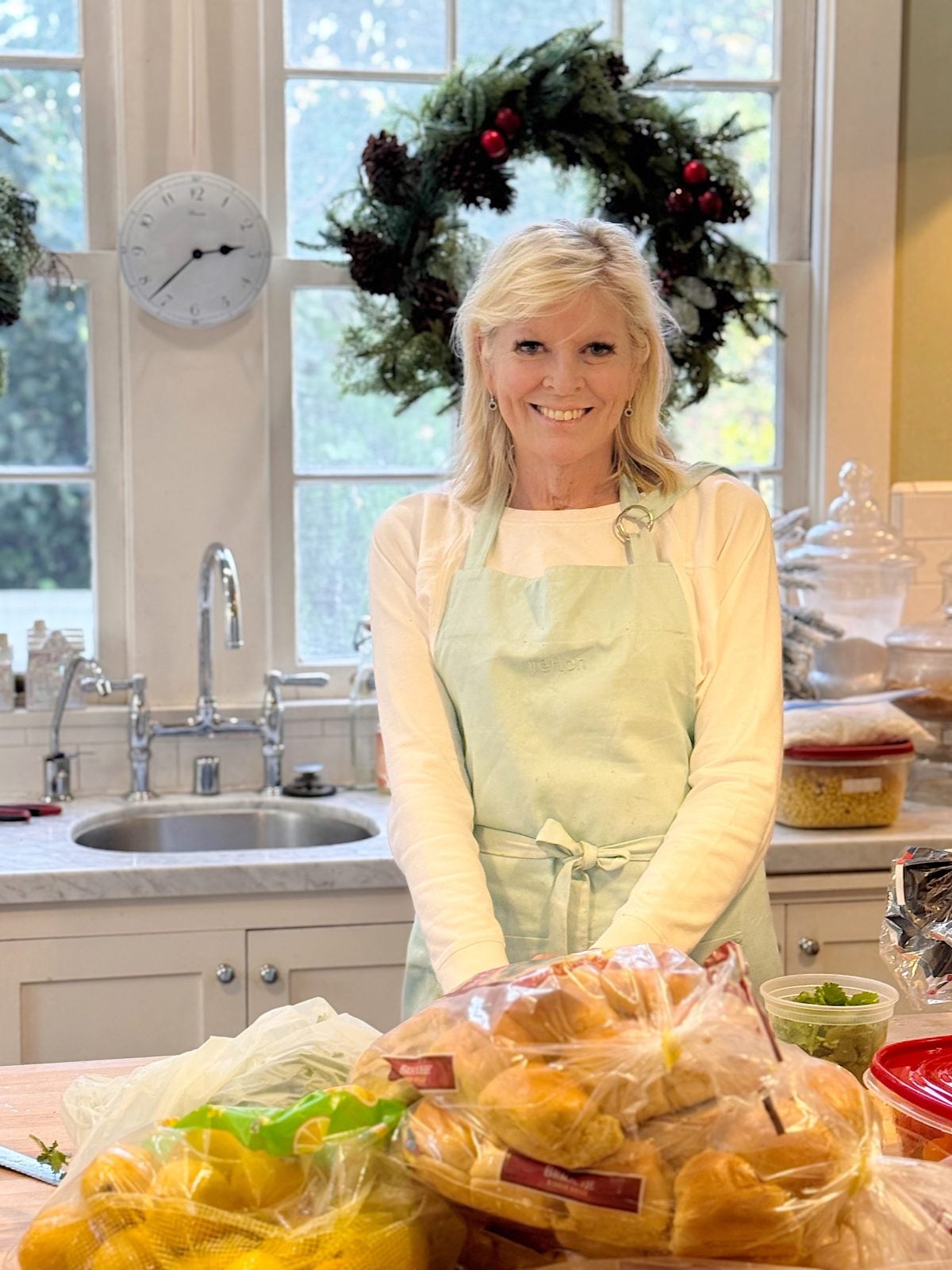 A woman wearing a light green apron stands in a kitchen with bags of bread rolls on the counter; a window with a holiday wreath is in the background.