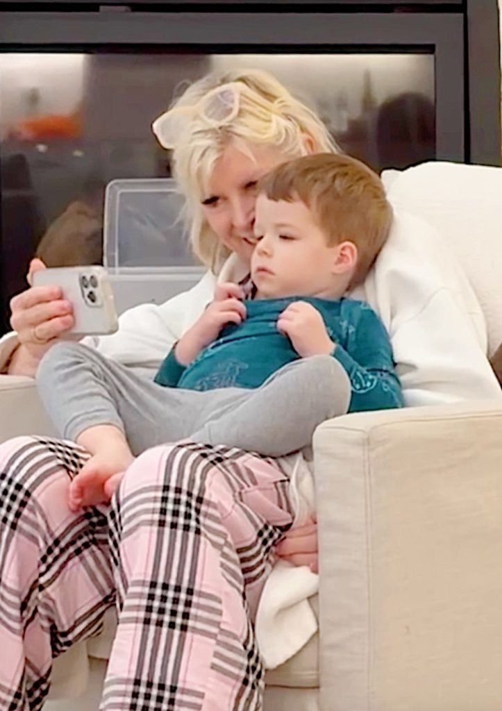 An adult and a child sit on a couch looking at a smartphone together; the adult wears pink plaid pants and the child wears grey pants and a teal shirt.