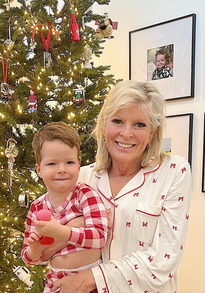 An older woman and a young boy in matching pajamas stand in front of a decorated Christmas tree, both smiling. Framed photos are visible on the wall in the background.