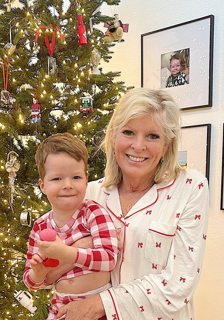 An older woman and a young boy in matching pajamas stand in front of a decorated Christmas tree, both smiling. Framed photos are visible on the wall in the background.