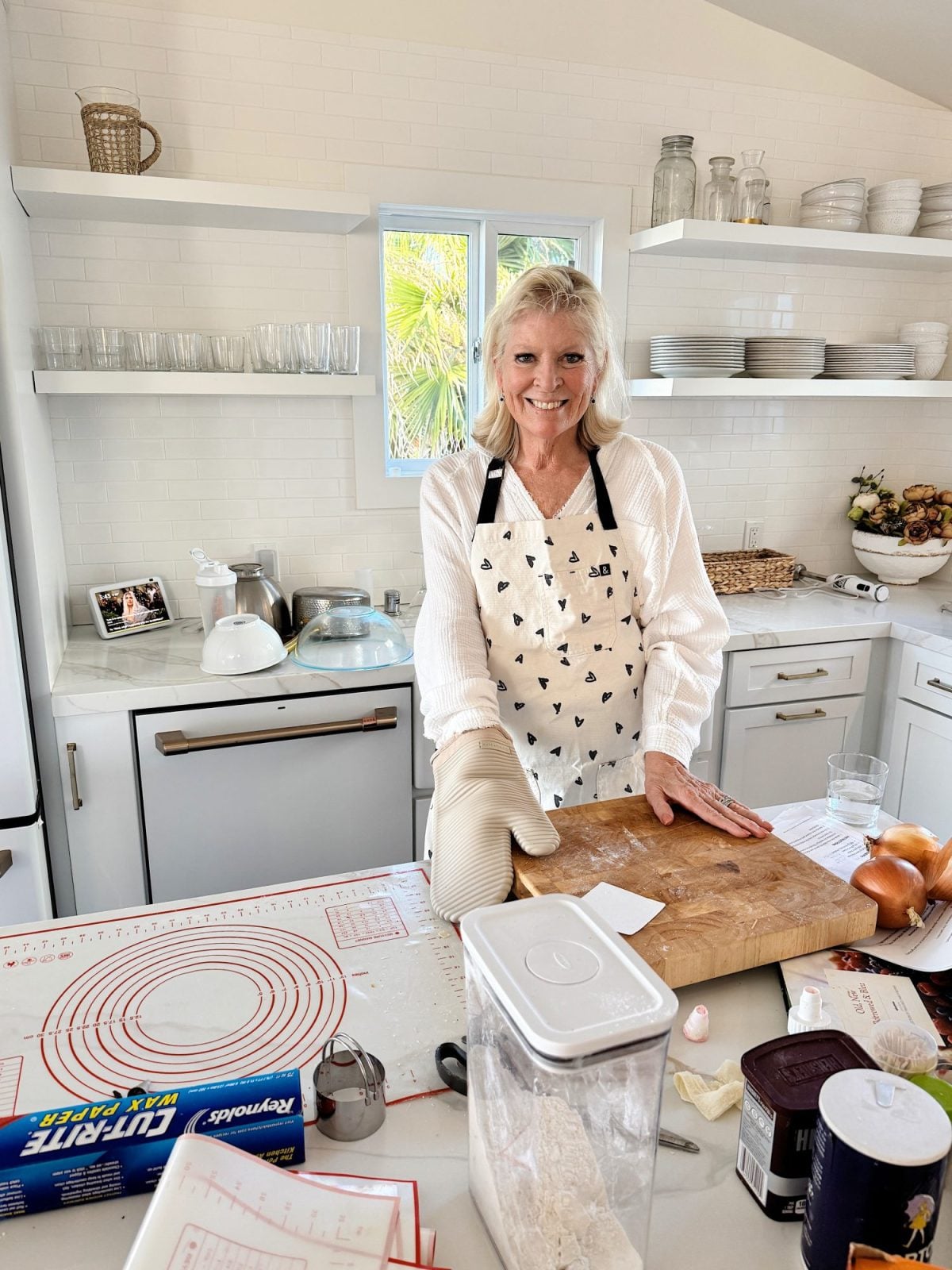 A woman wearing an apron and oven mitt stands in a bright kitchen, smiling while preparing food on a wooden cutting board with baking supplies around her, ready for hosting Christmas.