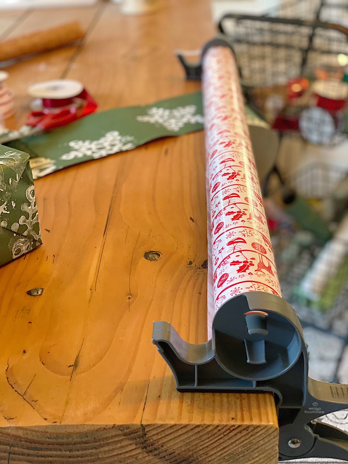 A roll of festive wrapping paper is placed on a wooden table, with a green wrapped gift and other wrapping supplies in the background.