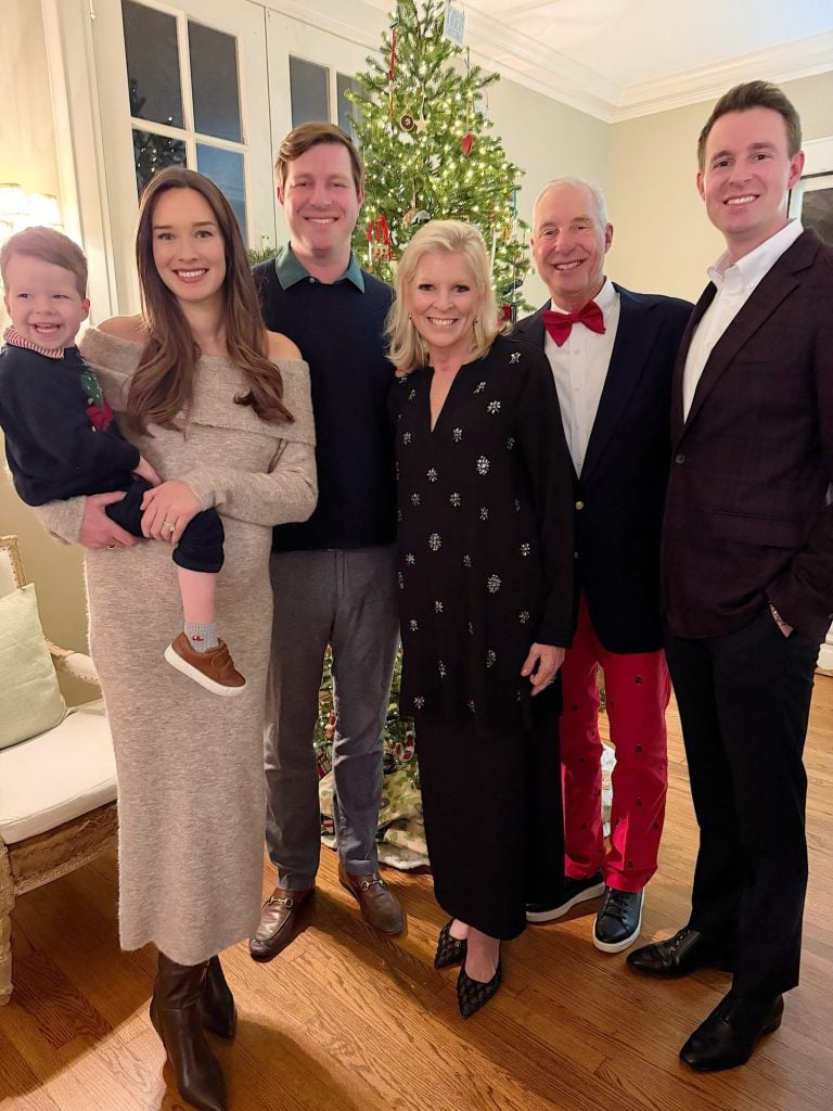 Six adults and a young child stand smiling in front of a decorated Christmas tree in a living room.