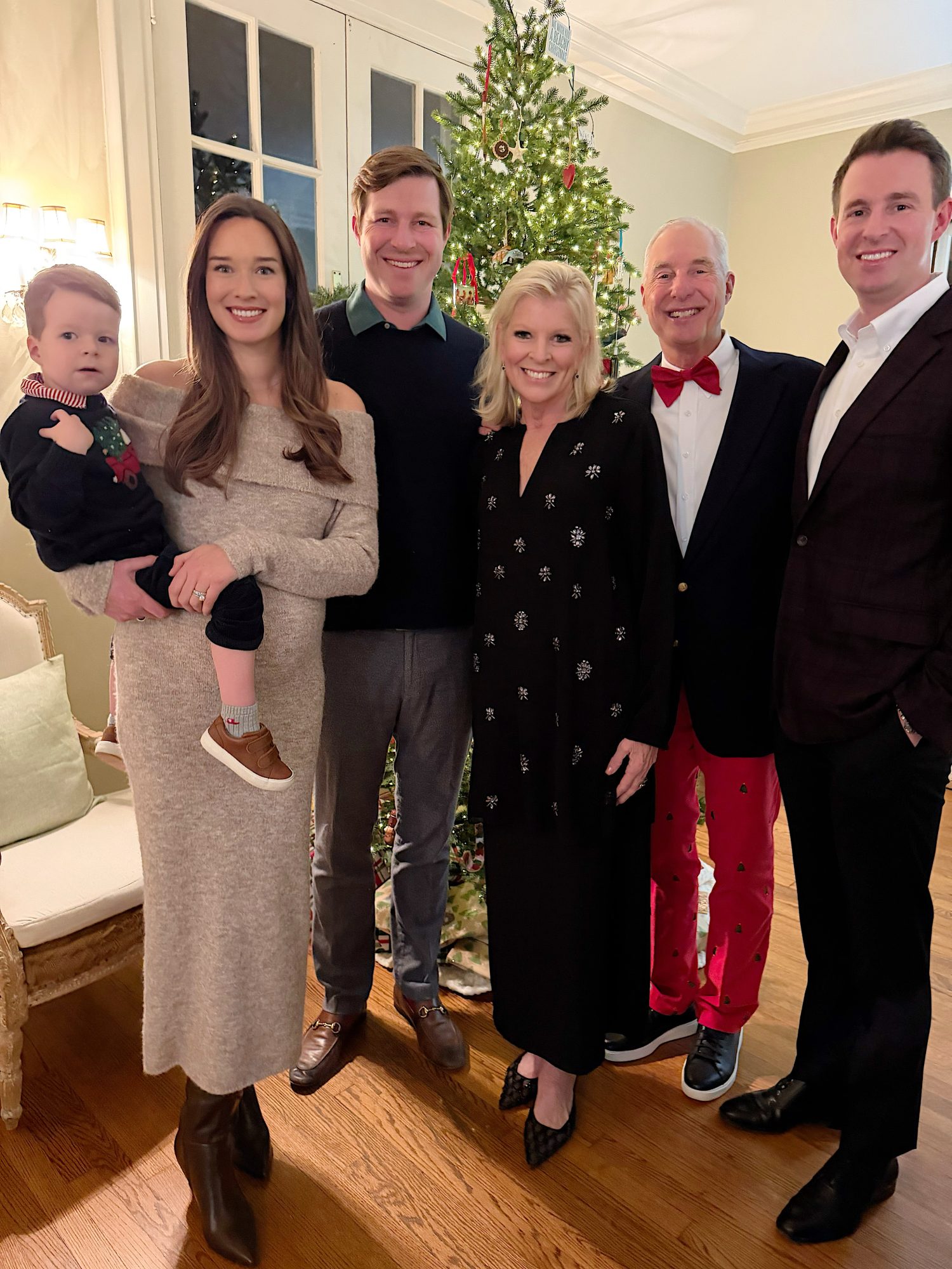 Six adults and one small child stand smiling in front of a decorated Christmas tree in a warmly lit living room.