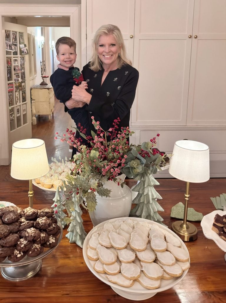 A woman holding a child stands behind a table with various cookies, greenery arrangements, and two small lamps in a well-lit room.