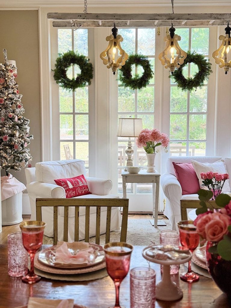 A living room decorated for Christmas with wreaths on French doors, a flocked tree, pink and white accents, and a dining table set with pink glassware and floral centerpieces.