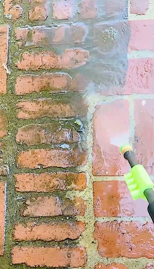 A person uses a pressure washer, an Amazon Find, to clean dirt and grime off a brick wall, revealing the brighter color of the clean bricks.