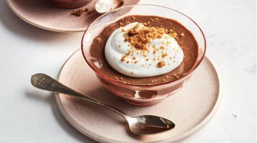 A glass bowl of chocolate mousse topped with whipped cream and crumbled cookies on a plate with a spoon beside it.