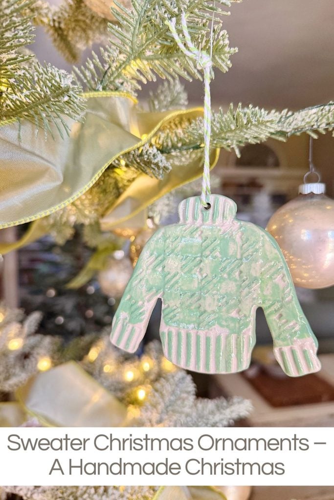 A handmade, green sweater-shaped ornament hangs on a Christmas tree next to a round white ornament and ribbon.