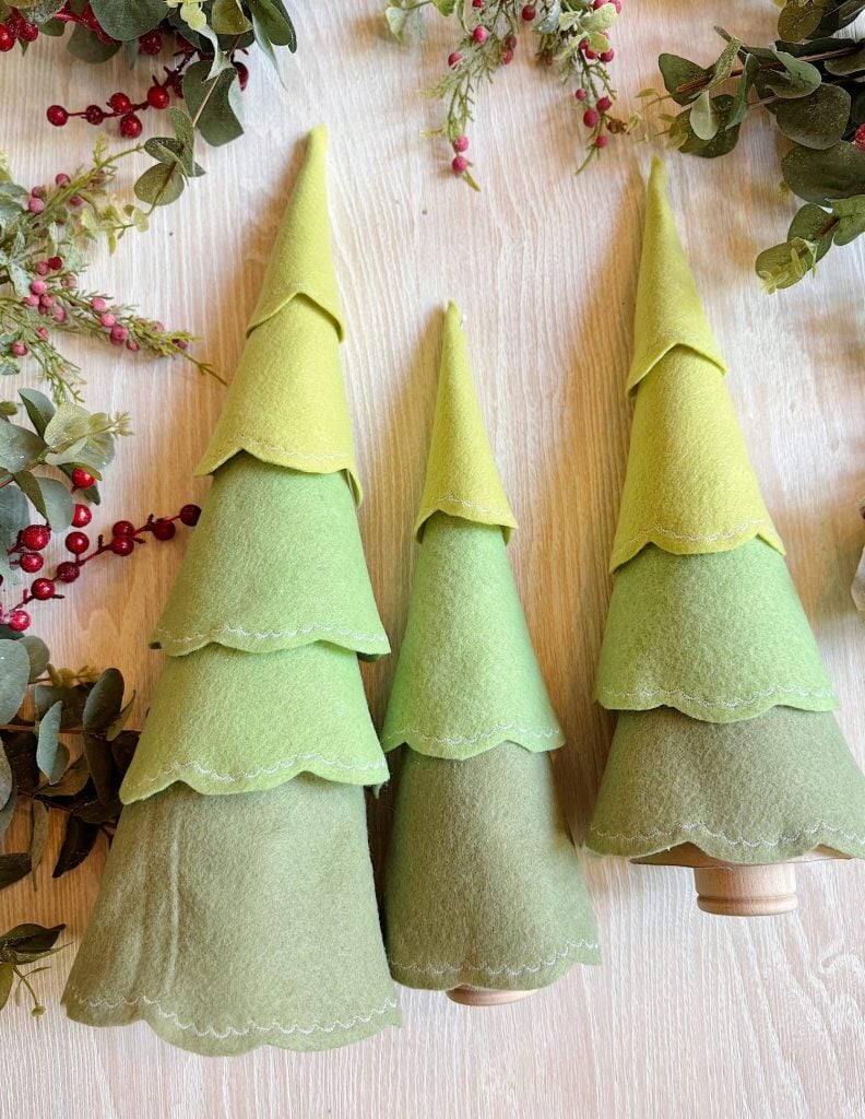 Three cone-shaped felt Christmas trees in green shades are displayed on a light wooden surface, surrounded by artificial greenery and red berries.