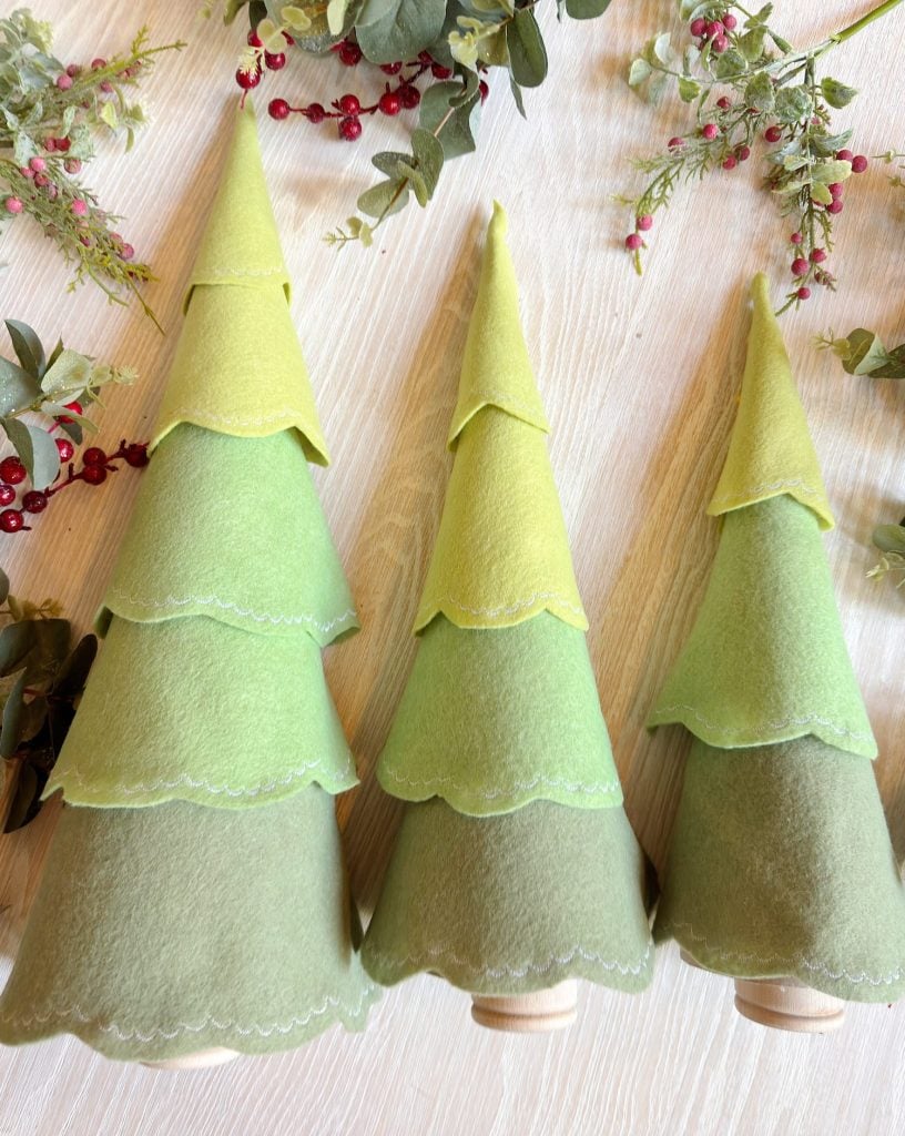 Three green felt cone trees with scalloped edges stand on a light wood surface, surrounded by sprigs of faux leaves and red berries.