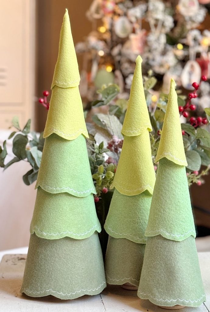 Three cone-shaped felt Christmas trees in varying heights and shades of green are displayed on a table with holiday decorations in the background.