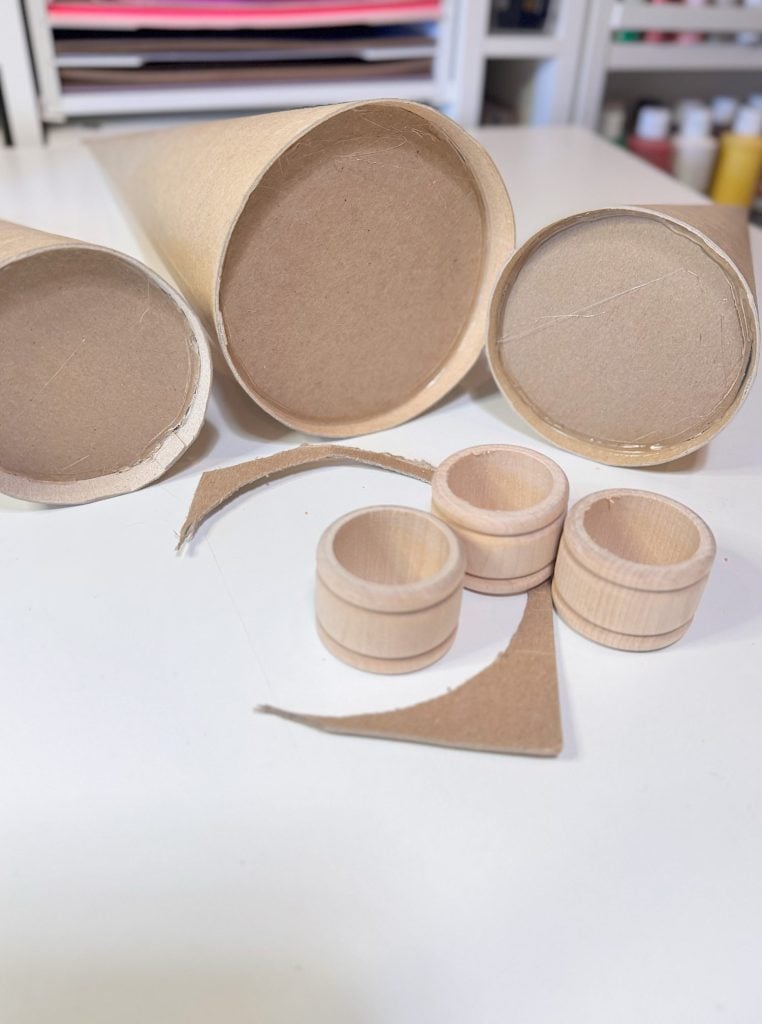 Three cardboard tubes, several cut cardboard rings, and three small wooden rings are displayed on a white surface, with additional craft supplies in the background.