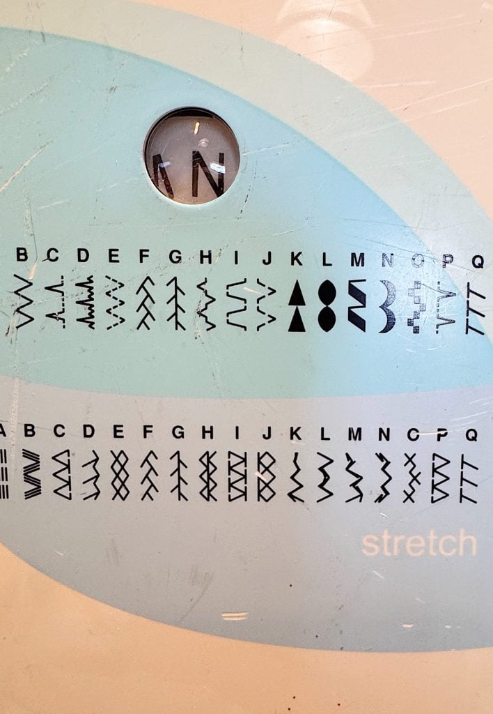 Close-up of a sewing machine stitch selector showing various stitch pattern options labeled with letters A to Q and the word "stretch" on the lower right.