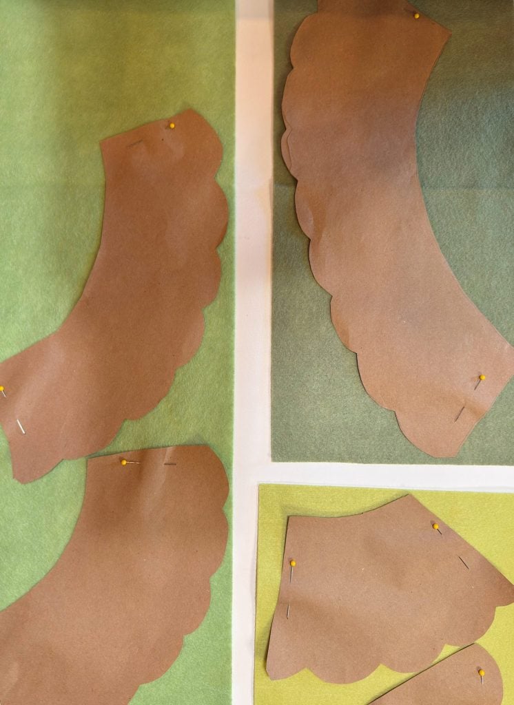 Four brown scalloped paper patterns are pinned to green felt fabric squares, arranged on a flat white surface.