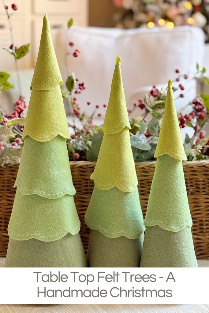 Three green felt tabletop Christmas trees of varying heights are displayed in front of a wicker basket decorated with festive holiday accents.