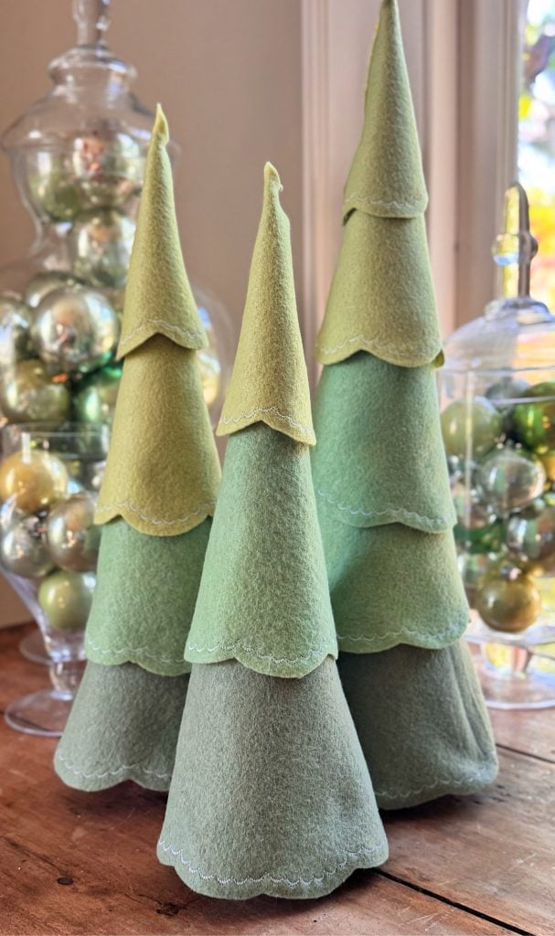 Three fabric Christmas trees in shades of green are displayed on a wooden surface, with glass jars of green and silver ornaments in the background.
