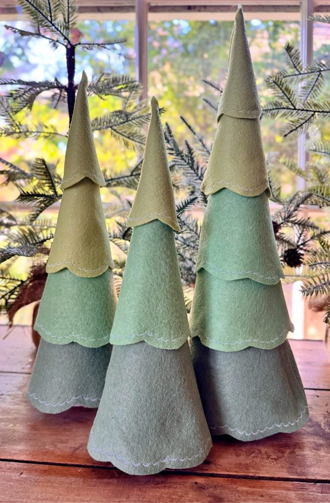 Three cone-shaped felt trees in varying shades of green are displayed on a wooden surface, with artificial pine branches and a window in the background.
