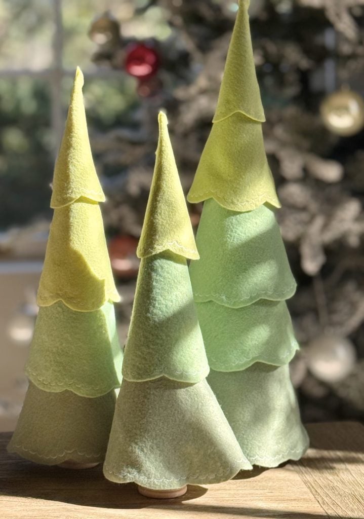Three pastel green and yellow felt Christmas tree decorations sit on a wooden surface, with a blurred decorated Christmas tree in the background.