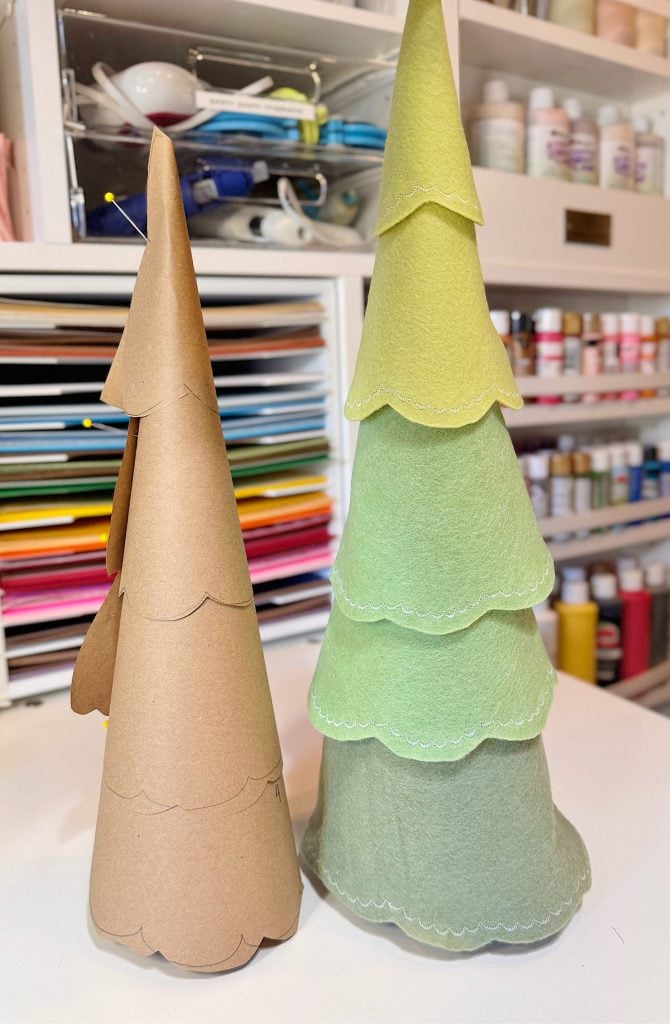 Two cone-shaped trees, one covered in brown paper and the other in layered green felt, sit on a craft table with colorful paper and art supplies in the background.