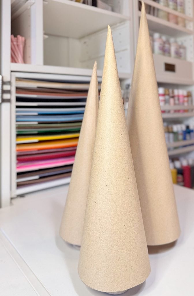 Three plain, cone-shaped cardboard forms are standing on a white surface in front of shelves filled with colored paper and craft supplies.