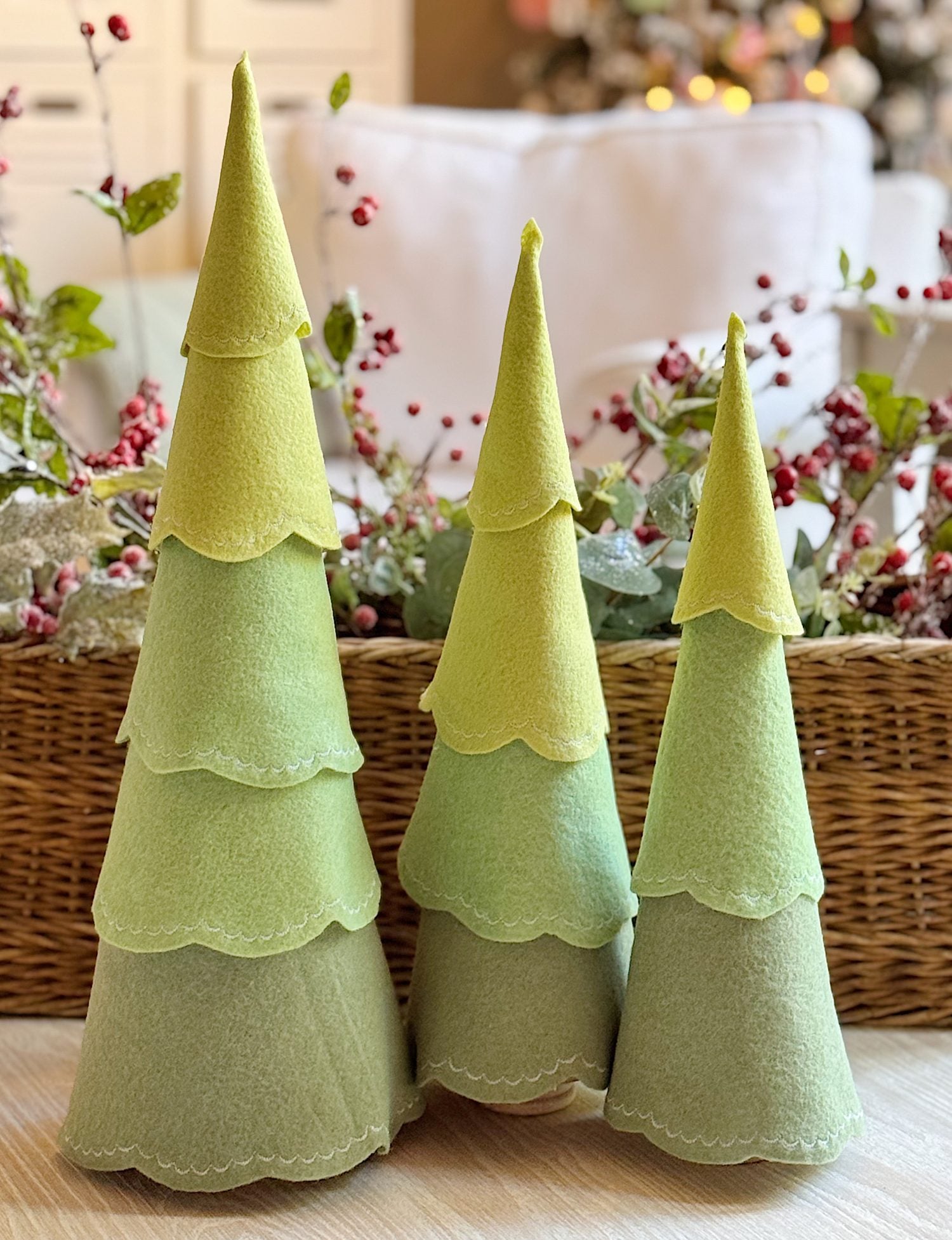 Three cone-shaped felt Christmas trees in varying shades of green are displayed in front of a woven basket with festive decorations and greenery in the background.