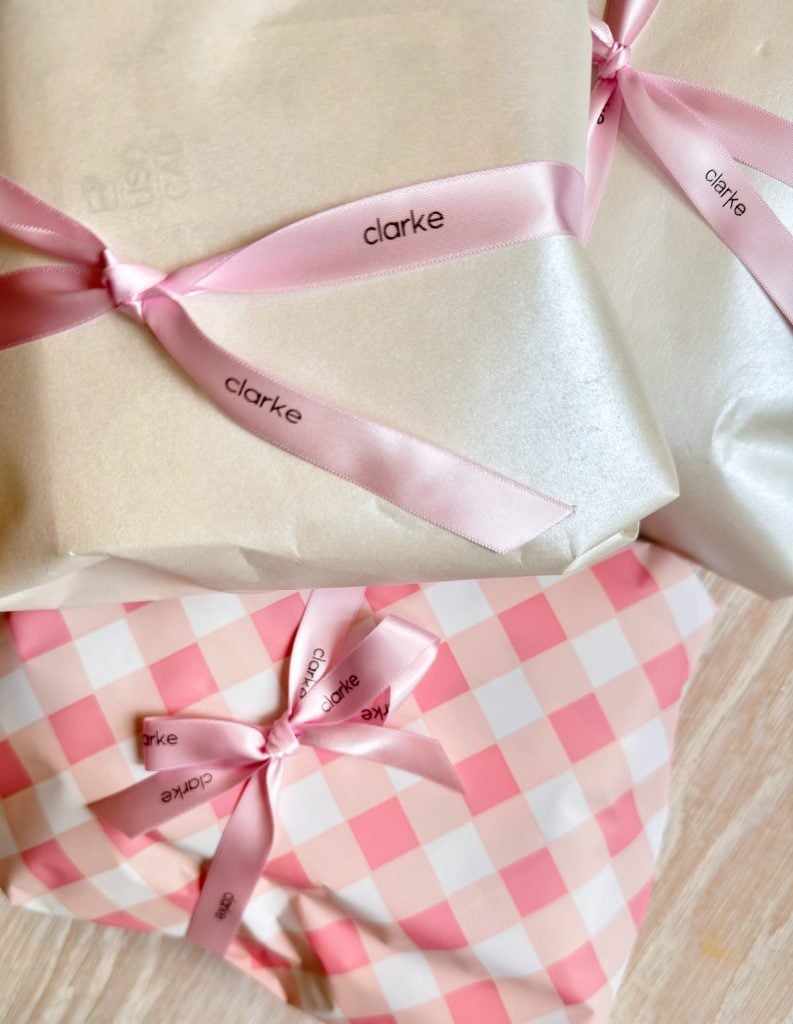 Three wrapped gifts, two in white paper and one in pink gingham, all tied with pink ribbons printed with the name "clarke.