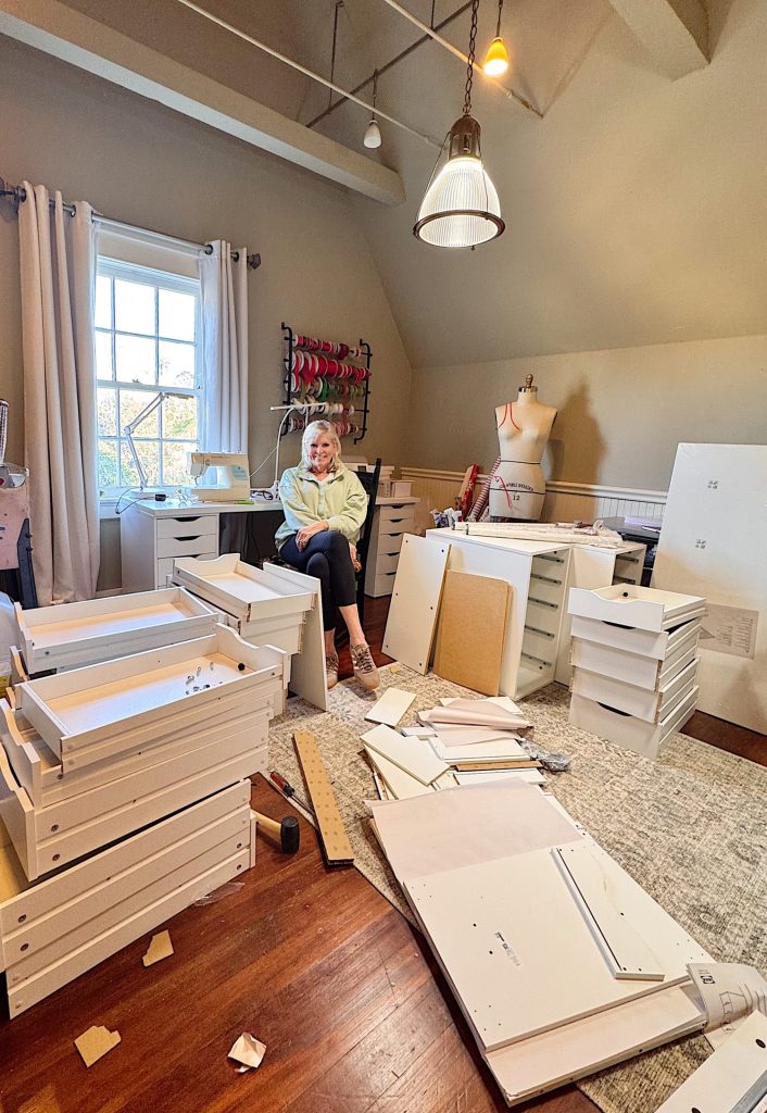 A woman sits on a chair in a room surrounded by unassembled white furniture pieces, cardboard, and tools, with a dress form and shelves in the background.