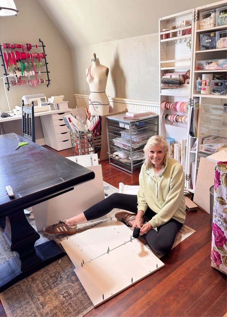 An older woman sits on the floor assembling a piece of white furniture in a craft room filled with organized supplies, a table, and a dress form.