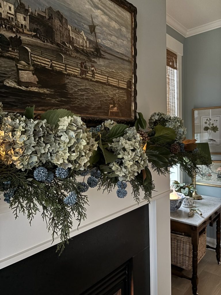 A mantel decorated with a garland of hydrangeas, greenery, and blue accents beneath a framed painting; a candle and framed art sit on a side table nearby.