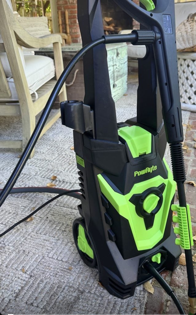 A green and black PowRyte pressure washer is positioned on an outdoor rug, surrounded by patio furniture and fallen leaves.